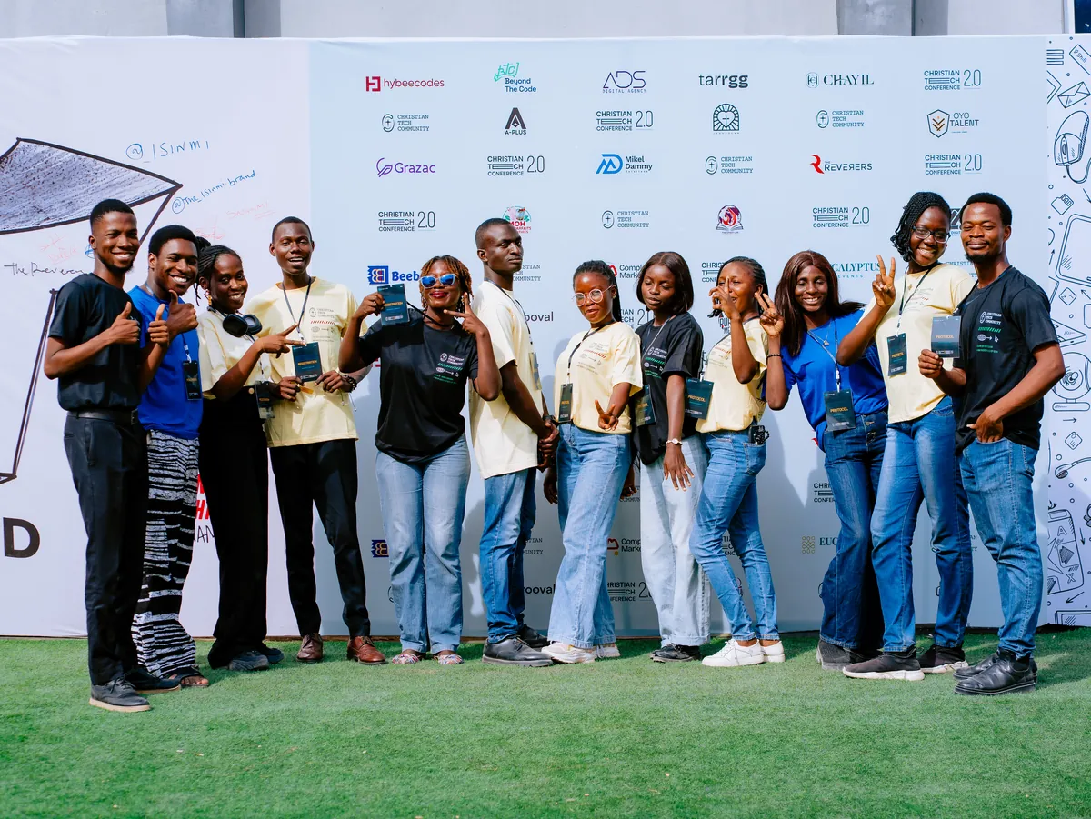 A group of about fifteen people in matching event shirts pose together on grass in front of a sponsor wall at CTC 2.0.